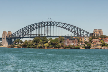 Sydney, Australia - December 11, 2009: Harbour Bridge, full metal span, bow and stone anchor towers against blue sky and above East shore with green vegetation and buildings. Azure bay in front.