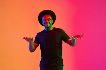 Poster Young african-american musician singing on gradient orange-purple studio background in neon light. Concept of music, hobby, festival. Joyful party host, stand upper. Colorful portrait of artist. © master1305