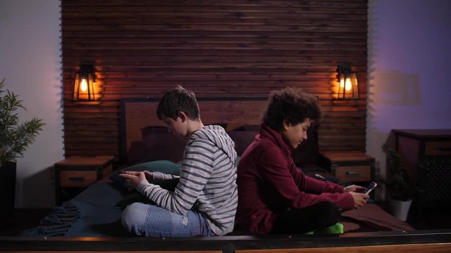 Indifferent mixed race friends phubbing each other while chatting online on smartphones at home. Addicted with cellphones diverse teenage boys spending joint free time serfing the net apart