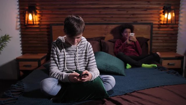 Addicted with gadgets mixed race teenage friends spending joint leisure chatting on smartphones apart in room. Diverse boys phubbing each other while communicating online on cellphones at home