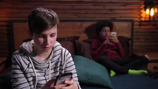 Close-up of diverse teenagers addicted with gadgets during free time at home. Focused on smartphones teenage boys phubbing each other while chatting online sitting on bed apart