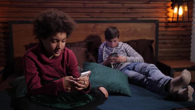 Close-up of busy on cellphones mixed race teenage friends during joint leisure in domestic room. Teenage schoolboys ignoring each other sitting on bed apart and surfing the net on smartphones