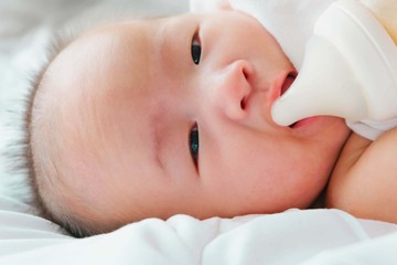 Portrait of a newborn Asian baby on the bed drinks milk from a bottle, Charming black-eyed baby 4 month old lies in bed ,A child resting on a bed