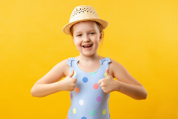 Happy cute beautiful charming little girl in a straw hat and a swimsuit shows thumb up with two hands. Close-up portrait of a child on a yellow background