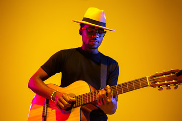 Obraz premium Young and joyful african-american musician playing guitar and singing on gradient orange-yellow studio background in neon light. Concept of music, hobby, festival. Colorful portrait of modern artist.
