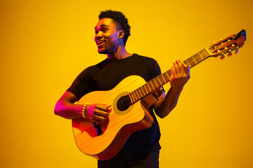 Obraz na płótnie Canvas Young and joyful african-american musician playing guitar and singing on gradient orange-yellow studio background in neon light. Concept of music, hobby, festival. Colorful portrait of modern artist.
