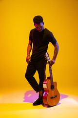Young and joyful african-american musician playing guitar and singing on gradient orange-yellow...