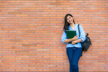 Asian smiling woman student holding book posing on brick background in campus. Happy teen girl high school student outdoors..