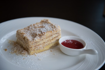 A piece of Napoleon cake on a white plate. The cake is sprinkled with powdered sugar. There is a Cup of jam next to it.