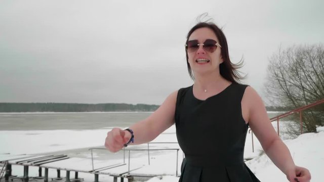 Woman in black dress and sunglasses jumps and dances on river bank covered with snow to make herself warm. Front view. Copy space.