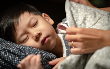 Closeup, cute Asian little boy sleep and mother's hand is cover him with blanket to keep him warm and get deep sleep at night. Mother's love and caring, Growth hormone and sleep connection concept.