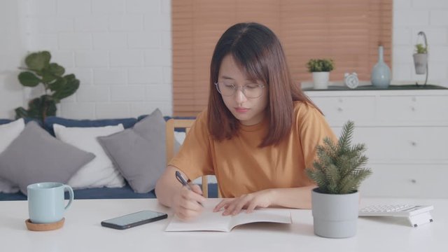 Attractive Asian Teenager Female College Student writing or making notes in a notebook doing homework exam or academic research sitting alone in living room at home.People and Education Concept