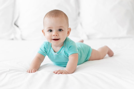 Happy Baby In Teal Onesie Lying On Tummy On White Bed