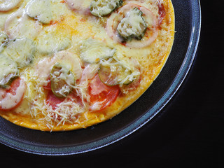 Hot pizza with tomatoes, grated Parmesan cheese, pickles and sauce in a glass plate on a dark background. Delicious homemade fast food recipe closeup.