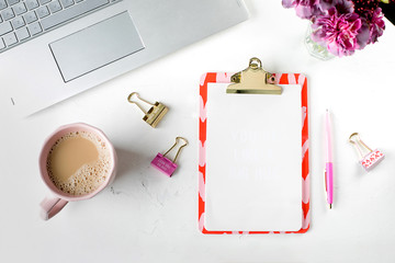 White fashion feminine home office workspace with laptop, pink flowers, cup of coffee, blank paper clipboard, pen and clips. Top view. copy space, flat lay. Mockup concept