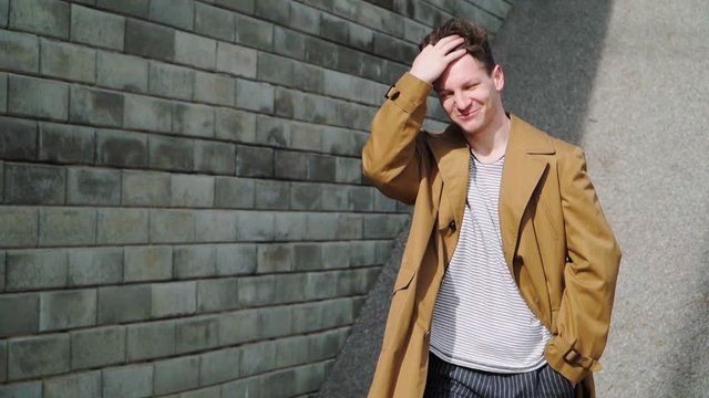 Stylish Beautiful Young Happy Caucasian Guy Goes along the City Wall and smiles. He is dressed in a Brown Long Coat. Slow motion. Close up - Powered by Adobe