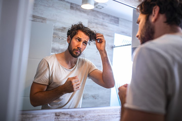 Handsome young man touching his hair with hand and grooming in bathroom at home. White metrosexual man worried for hair loss and looking at mirror his receding hairline.