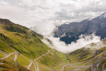 Mountain road zigzagging up green lush Dolomites surrounded by bright white moody clouds