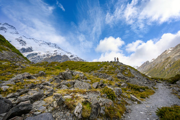 Two tourists stand on the hillside to take panoramic pictures of the Mueller Glacier at Kea Point in Mount Cook National Park, the rocky mountains and summer green grass in New Zealand.