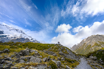 Two tourists stand on the hillside to take panoramic pictures of the Mueller Glacier at Kea Point in Mount Cook National Park, the rocky mountains and summer green grass in New Zealand.