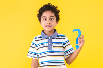 Curly pretty perplexed dark skinned boy in striped blue t-shirt is holding in hand paper question sign. Black cute child is smiling on yellow orange background. Emotional portrait concept.