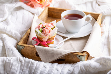 Flower cupcakes and cup of tea on wooden tray on white plaid.  Beautiful sponge cup cakes decorated with buttercream roses on white background.