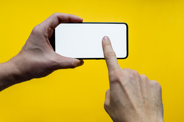 Male hands holding and touching the empty black smatphone screen in a horizontal position, isolated on a yellow background