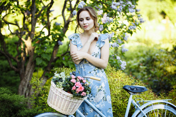 portrait of a beautiful girl in the forest, holding a bike with a basket of flowers, behind the rays of the sun, a blue flowered dress, summer walk