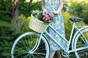 Fototapeta na wymiar portrait of a beautiful girl in the forest, holding a bike with a basket of flowers, behind the rays of the sun, a blue flowered dress, summer walk