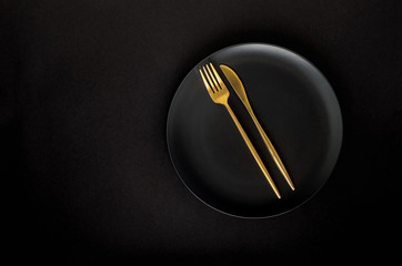 Black empty ceramic plate with golden cutlery (fork and knife) on a black background (table).