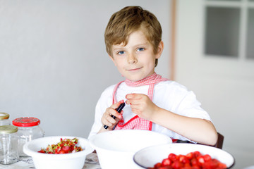 Healthy little blond kid boy helping and making strawberry jam in summer. Funny child cleaning berries and preparing for cooking jam. Kid eating ripe strawberries in domestic kitchen.