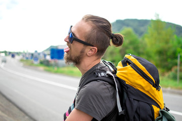 Young man in trendy standing in the middle of highway and looking away. Full-length portrait of traveler with big backpack walking down the road in morning. Emotional traveler in adventure.