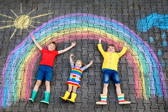 three little children, two school kids boys and toddler girl having fun with with rainbow picture drawing with colorful chalks on asphalt. Siblings in rubber boots painting on ground playing together.