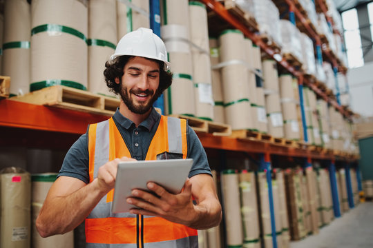 Male Warehouse Manager Adding Stock Inventory Data In Digital Tablet In Warehouse Wearing A White Hardhat And Safety Vest Standing