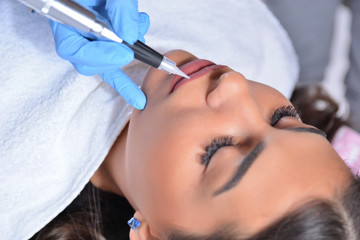 Young pretty girl having permanent makeup on lips in beautician salon. Cosmetologist applying tattoo with the professional and sterile tool. Close up, selective focus