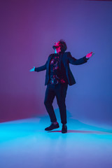 Obraz premium Young caucasian musician in casual dancing on gradient blue-purple background in neon light. Concept of music, hobby, festival. Joyful party host, DJ, stand upper, dancer. Colorful portrait of artist.