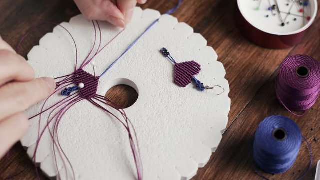 Lifestyle concept, work from home to reinvent your life: close-up of woman hands making macrame knots the fuchsia thread creating an earring on kumihimo tools on wooden table