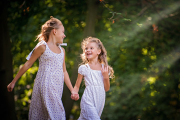 Obraz premium Two curly blonde girls in dresses running along the path of a summer park and laughing