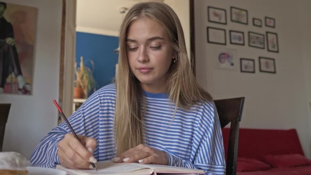 A focused nice blonde young woman is witting something in her notebook while sitting in the living room
