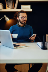 Handsome bearded caucasian hipster sitting in his modern home office, using smart phone for checking messages on social media and holding cup of coffee.