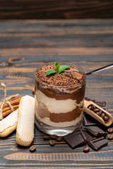 Classic tiramisu dessert in a glass cup, pieces of chocolate and savoiardi cookies on wooden background