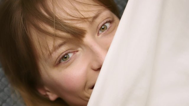 Happy young woman playfully hiding under sheets. Close up slow motion shot. joyful facial expression.