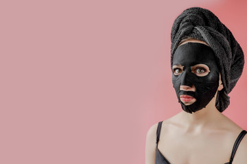 Young woman apply black cosmetic fabric facial mask on pink background. Face peeling mask with charcoal, spa beauty treatment, skincare, cosmetology. Close up
