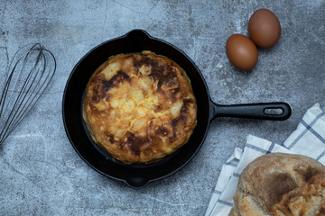 Potato and egg omelette in a pan, homemade bread on a cloth, two raw eggs and a whisk.