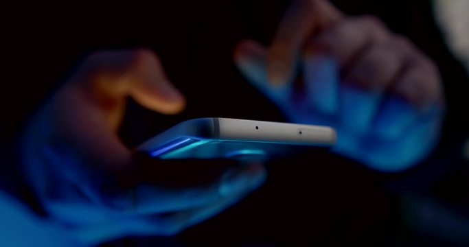 very close-up of the bottom and end parts of the smartphone. men's hands are out of focus and holding the phone. the fingers slide across the screen. lower blue light