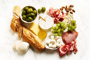 Cheese plate with brie, parmesan, cheddar and meat. Flatlay with variety of gourmet snacks, fruits and baguette on marble board.