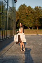 Beautiful smiling stylish girl wearing white fashionable dress walking with shopping bags.