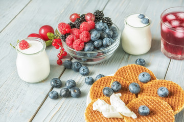 Different types of ripe berries - blueberries, raspberries, red currants, mulberries, a drink from berries and waffles with cheese. Appetizing snack with berries. Jars of natural yogurt.