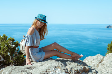 A young woman in a summer dress sits on the edge of a mountain or cliff with a beautiful sea view
