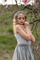  young blonde in a gray dress with a long train in the garden where pink magnolia blooms in spring, close-uo